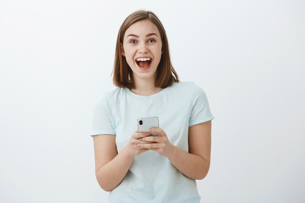 amazed and excited attractive young european female model smiling broadly from great news received via internet holding smartphone gazing thrilled and delighted posing against white wall