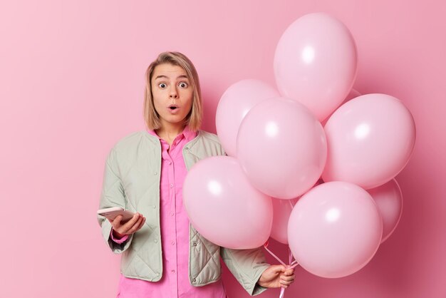 Amazed faired haired young woman stares embarrassed reacts on shocking news wears shirt and jacket poses with inflated of balloons and smartphone isolated over pink background Holidays concept