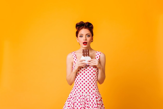 Foto gratuita modello femminile stupito in vestito a pois che guarda l'obbiettivo. studio shot di pinup donna con i capelli allo zenzero tenendo il cioccolato.