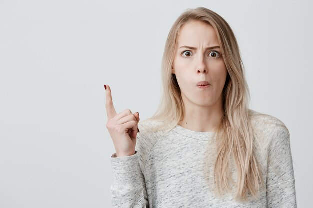 Amazed female model with straight dyed blonde hair, wearing casual clothes, pointing with forefinger at copy space