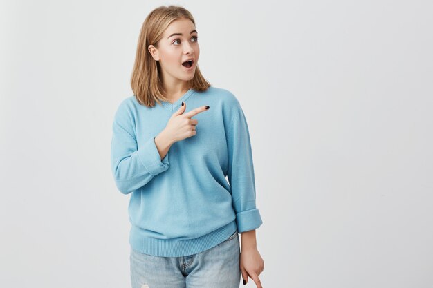 Amazed female model with straight fair hair, wearing blue clothes, looking with bugged eyes and widely opened mouth, pointing with forefinger at copy space for your text or advertisment