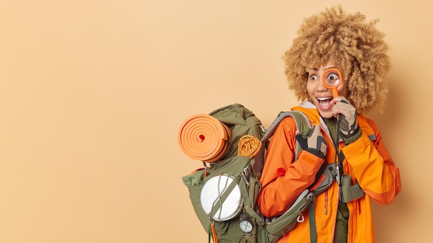 Amazed female tourist looks through magnifing glass dressed casually carries rucsack with rolled kareamt binoculars on neck isolated over brown background mock up space for your advertisement