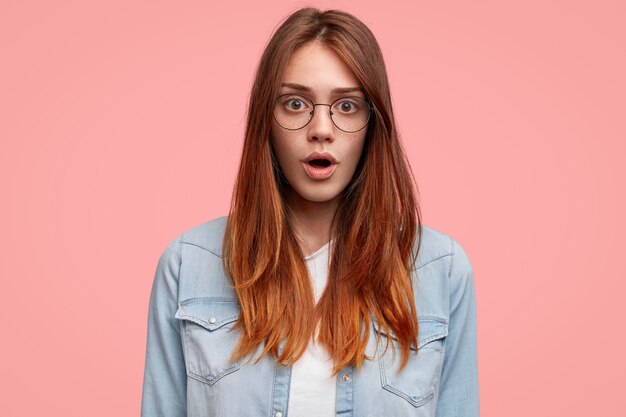 Amazed freckled female with wide opened mouth, has straight hair and freckled skin, wears round spectacles and fashionable denim jacket, being afraid of something.