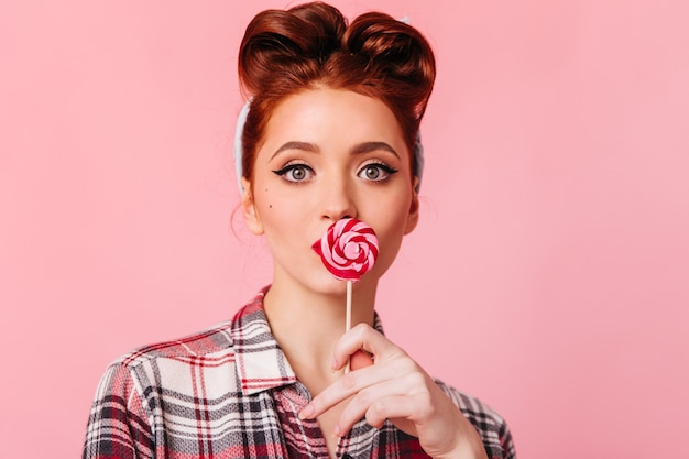 Free photo amazed ginger girl in checkered shirt looking at camera. studio shot of pinup woman with candy isolated on pink space.