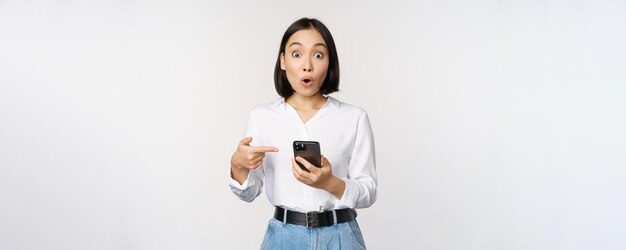 Amazed girl talking about smartphone app pointing at phone while looking impressed at camera standin