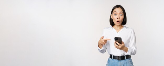 Amazed girl talking about smartphone app pointing at phone while looking impressed at camera standing against white background