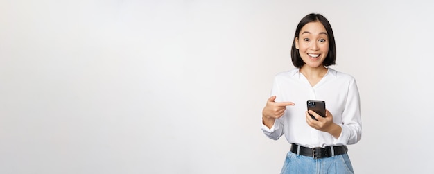 Amazed girl talking about smartphone app pointing at phone while looking impressed at camera standing against white background