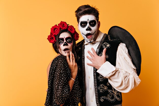Amazed guy and girl with painted faces for Halloween look in fright. Shot of couple in national Mexican costumes on isolated wall.