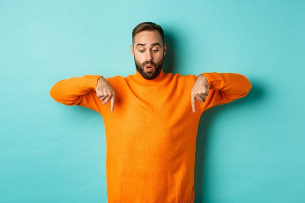 Amazed guy in orange sweater, pointing fingers down and looking with interest standing over turquoise wall.