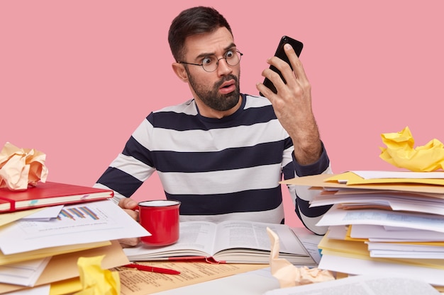 Amazed handsome man with thick stubble, wears transparent glasses, holds modern cell phone, recieves bad information