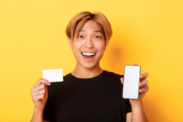 Amazed happy asian guy showing credit card and smartphone screen, smiling fascinated, standing yellow wall.