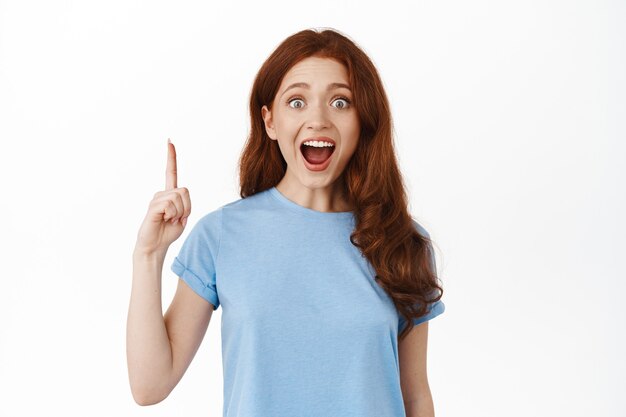 Amazed happy redhead girl showing copy space, pointing finger up and staring excited on white