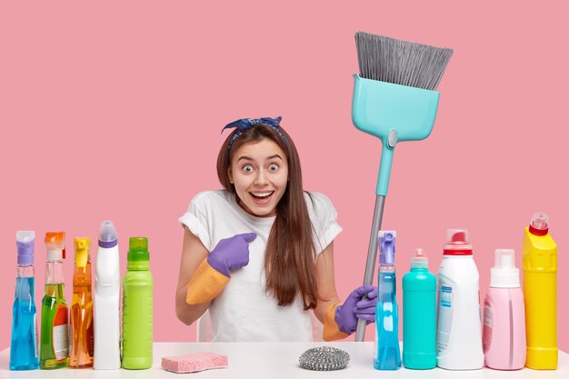 Amazed joyful young woman indicates at herself, works in cleaning service, holds broom, sits at desk with washing detergernts and deodorizer