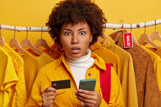 Amazed lady with Afro hairstyle, dressed in yellow shirt, poses over clothing racks, holds modern cellular and credit card