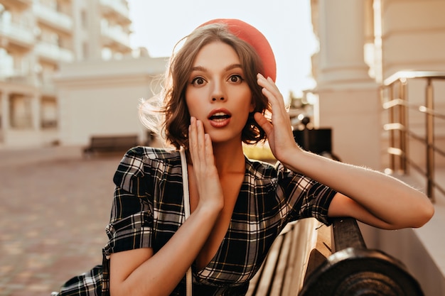 Amazed lady with short hair sitting on bench in warm day. Outdoor photo of attractive curly surprised girl in red beret.