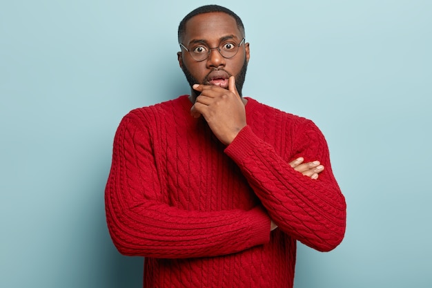 Amazed male employee holds chin, stares in disbelief, has embarrassed facial expression, problems at work, wears round glasses and red jumper. People and facial expressions.