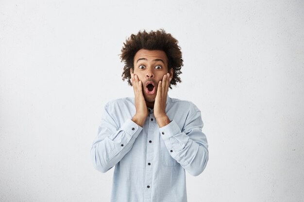 Amazed mixed race male with Afro haircut touching his cheeks looking with bugged eyes