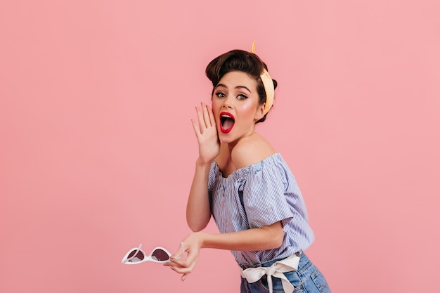 Signora pinup stupita in posa emotivamente su sfondo rosa. studio shot di meravigliosa ragazza con occhiali da sole.