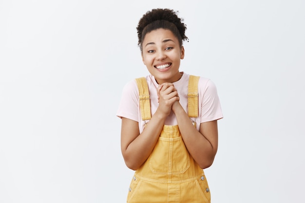 Amazed and pleased thrilled dark-skinned girl asking for autograph with clenched palms on chest, begging or asking celebrity with broad loving smile, standing friendly and excited over gray wall