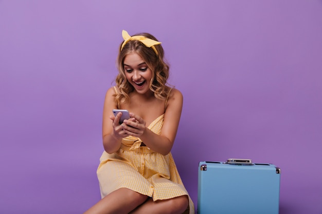 Бесплатное фото Пораженная красивая девушка с чемоданом, читая телефонное сообщение. портрет радостной кудрявой дамы с синим чемоданом, смотрящей на ее смартфон.
