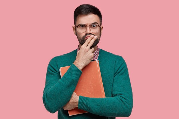 Amazed puzzled European guy with thick bristle, purses lips, dressed in green jumper, big square spectacles, holds red old textbook