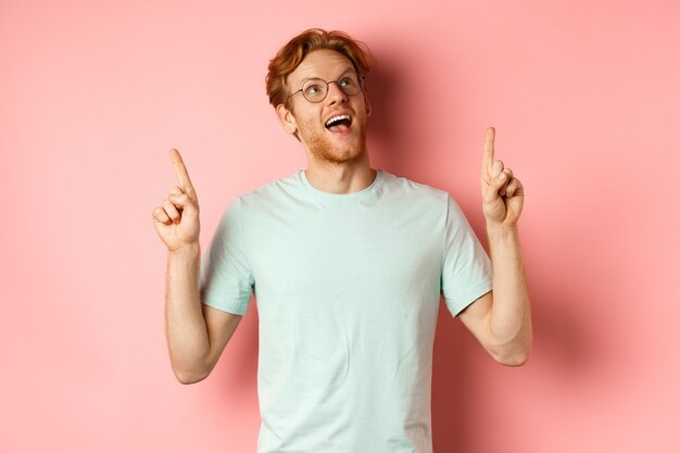 Amazed redhead guy in glasses checking out special deal looking and pointing fingers up with amazed ...