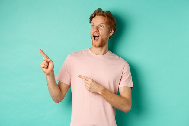 Amazed redhead man in t-shirt checking out promo, gasping in awe and pointing fingers at upper left corner, standing over mint background.