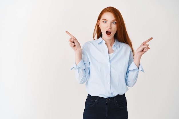 Amazed redhead woman manager pointing sideways, showing two variants and look surprised at front, white wall