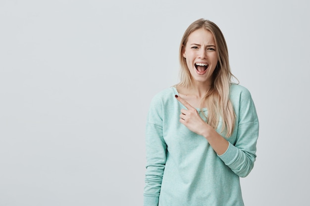 Amazed shocked female model with straight fair hair, wearing blue clothes, posing with widely opened mouth, pointing with forefinger at copy space