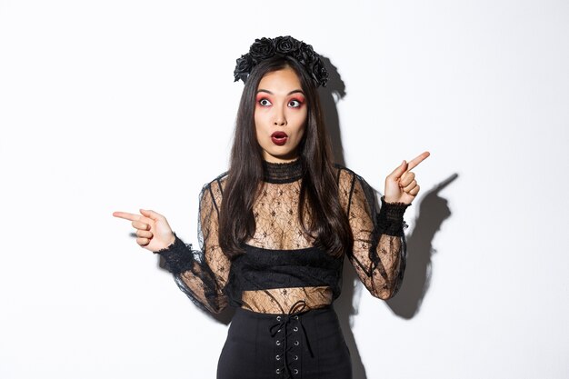 Amazed and startled woman in witch costume looking impressed, pointing fingers sideways but stare left, open mouth wondered, standing in black lace dress and wreath, white background.