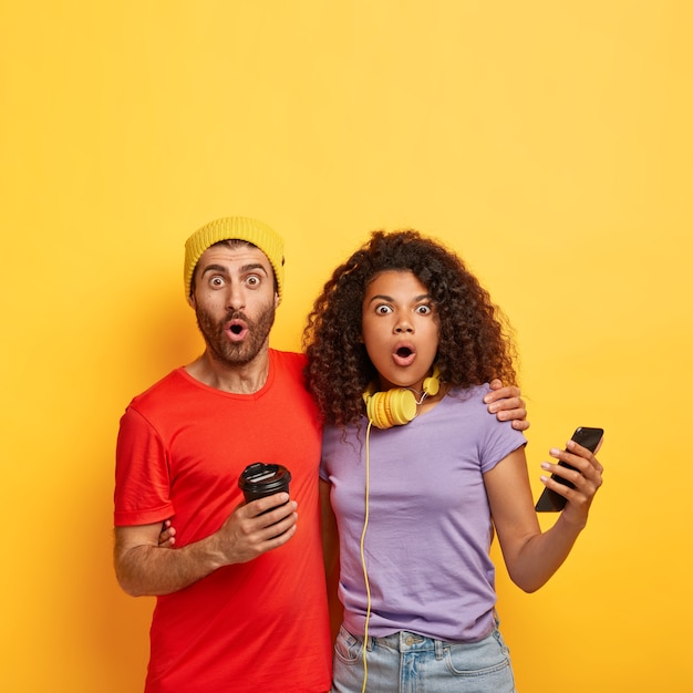 Amazed stylish couple posing against the yellow wall with gadgets