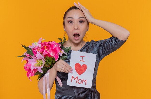 Amazed and surprised asian woman mother holding greeting card and bouquet of flowers celebrating mother's day standing over orange wall