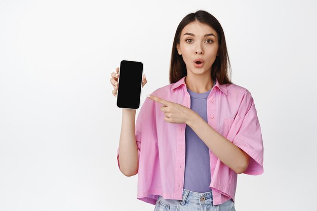 Amazed teen girl pointing finger at her mobile phone screen looking impressed discussing something on smartphone social media white background