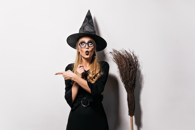 Amazed wizard in glasses standing on white wall. Funny emotional witch posing with hat and broom.