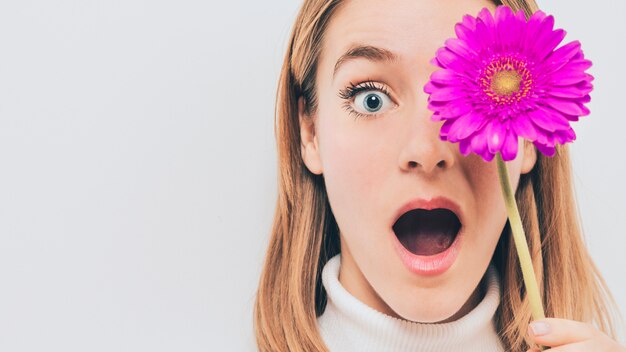 Amazed woman holding pink gerbera flower at face