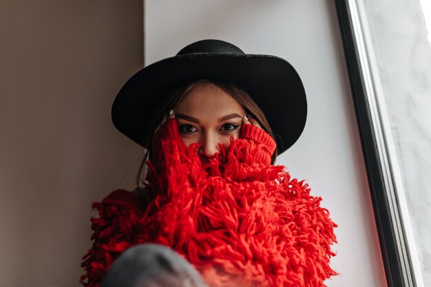 Amazed woman in red sweater and wide-brimmed black hat covers her face sitting on windowsill.