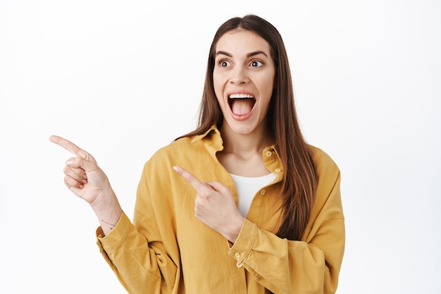 Amazed woman scream from joy and excitement, pointing and looking left with dropped jaw and fascinated face, stare in awe at logo promo banner, standing over white background.