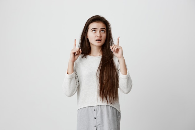 Amazed woman with straight long dark hair, wearing casual clothes, looking with dark eyes upwards and widely opened mouth, pointing with forefingers at copy space