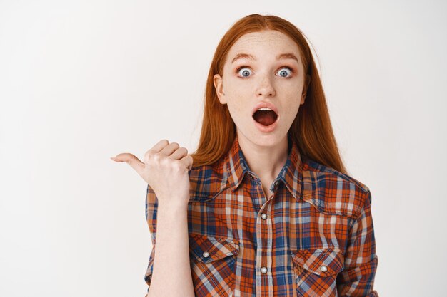 Amazed young female student checking out an advertisement, looking at at front with opened mouth, pointing finger left at promo banner, white wall