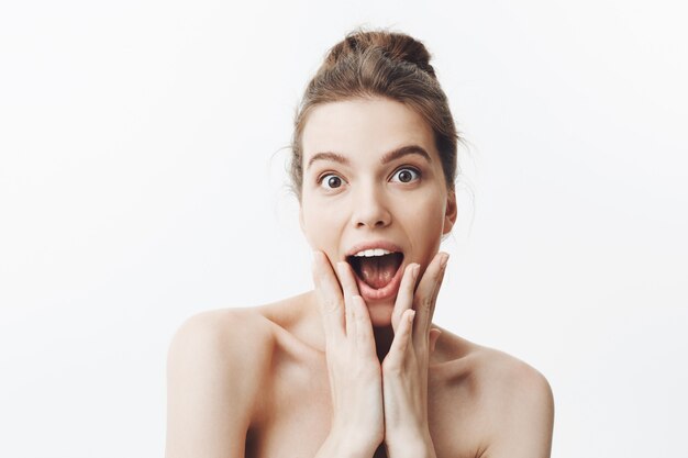 Amazed young good-looking european student girl with brown hair in bun hairstyle and naked shoulders, squeezing face with hands with surprised expression. Beauty and health