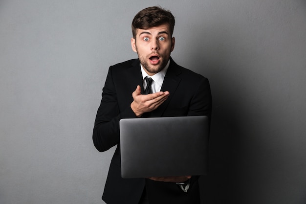 Amazed young guy in black suit holding laptop, 