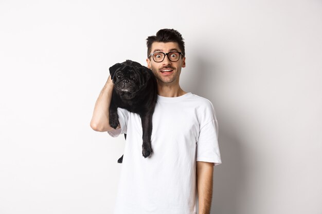 Amazed young man in glasses holding black pug on shoulder and staring at camera impressed. Dog owner posing with cute puppy near white background.