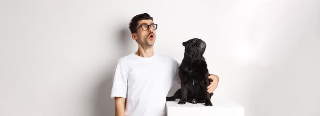 Free photo amazed young man in glasses hugging his dog pet owner and pug staring at upper left corner promo off