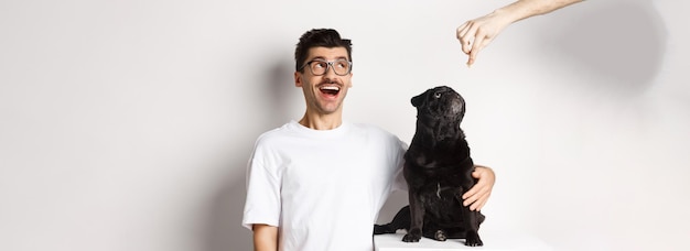 Изумленный молодой человек в очках, обнимающий владельца своего пса и мопса, смотрит на рекламу в верхнем левом углу