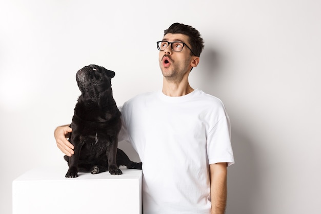 白い背景の上に立って、彼の犬、ペットの飼い主、左上隅のプロモーションオファーを見つめているパグを抱き締める眼鏡をかけた驚いた若い男。
