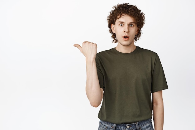 Amazed young man pointing left staring impressed at camera checking out advertisement showing promo offer store deal standing over white background