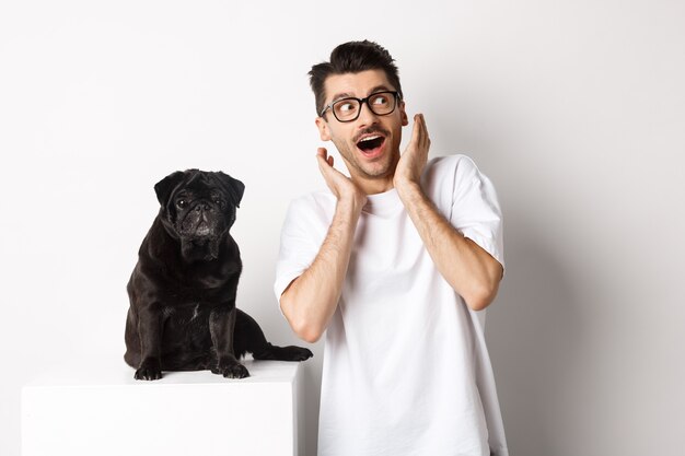Amazed young man standing with cute black puppy, staring at upper right corner surprised and excited, standing near pug over white background.