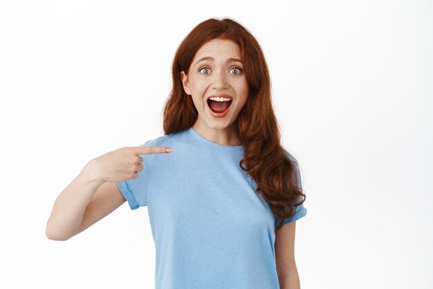 Amazed young natural woman, student with red hair in t-shirt pointing at herself with wondered and surprised face, being chosen, self-promoting, winning unexpectedly, white background.