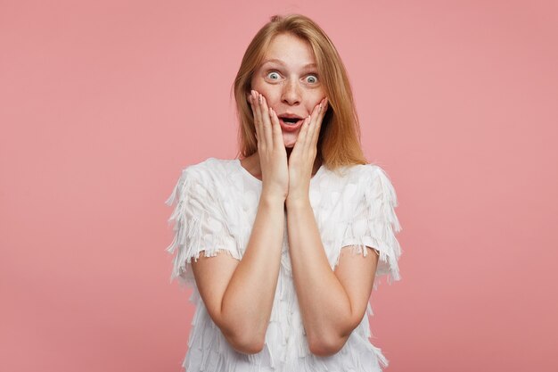 びっくりしたカメラを見て ピンクの背景の上にポーズをとって 上げられた手で彼女の顔を保ちながら 彼女の緑灰色の目を丸めるセクシーな髪の驚くべき若いきれいな女性 無料の写真