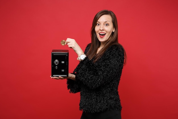 Amazed young woman in fur sweater holding metal bank safe for money accumulation, bitcoin, future currency isolated on red background. people sincere emotions, lifestyle concept. mock up copy space.